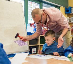 A teacher leans over a child, demonstrating how to use a paint stencil. Both are smiling