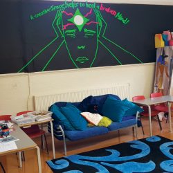 The room known as the Engine Room, containing a blue comfy sofa and rug, colouring things, and a board saying "a creative space helps to heal a broken mind"