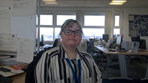 Katie in her office wearing a stripy shirt, looking at the camera with desks and windows behind