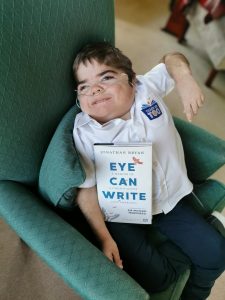 Jonathan Bryan, a Disabled Young campaigner and author is pictured with his book, 'Eye Can Write'. He is a Young white man wearing a shirt with the logo of his charity, 'Teach Us Too', on it.