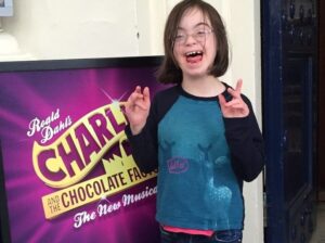 A photo of Sharon Smith's daughter smiling next to a sign for the Charlie and The Chocolate Factory Musical
